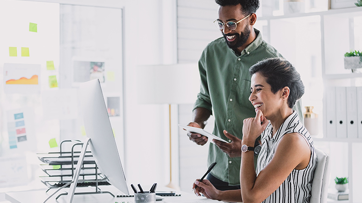 two co-workers smiling while looking at a monitor